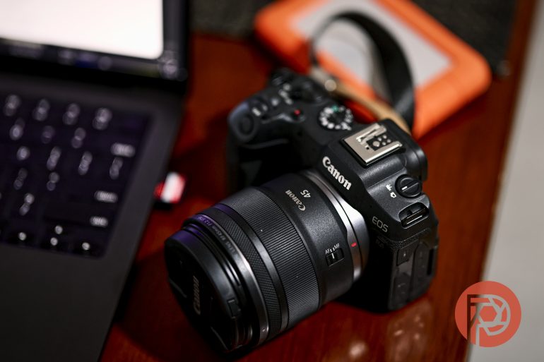 A Canon EOS camera and laptop on a wooden surface with an orange portable hard drive in the background.