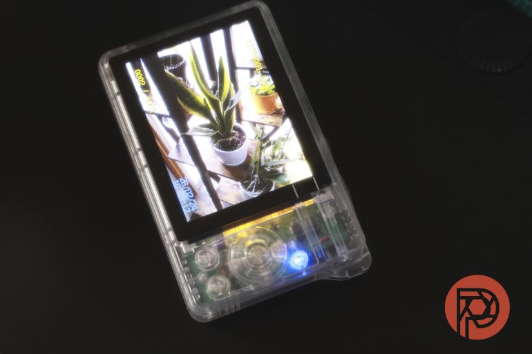A handheld game console with a clear shell displays a photo of potted plants on its screen.