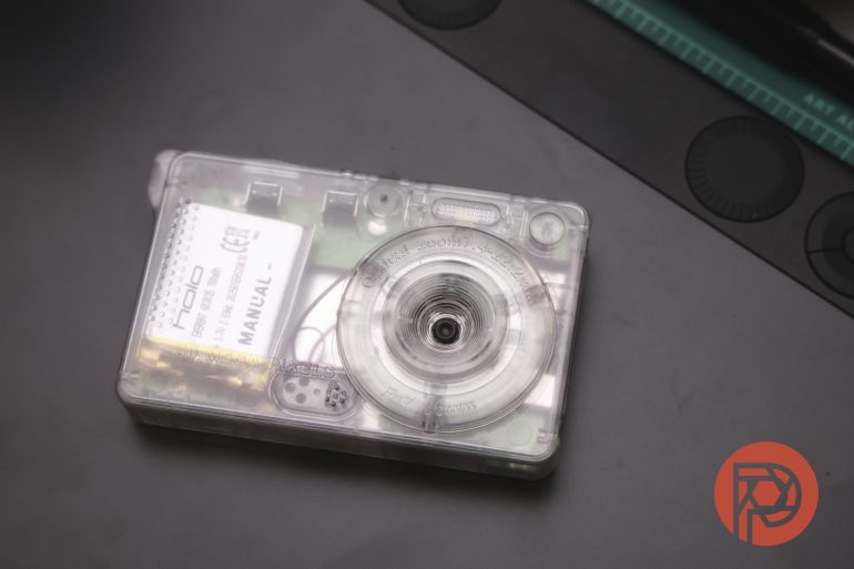 A transparent cassette tape with visible internal parts rests on a dark surface near a ruler.
