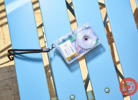 A small, transparent film camera with a wrist strap resting on a bright blue slatted bench.