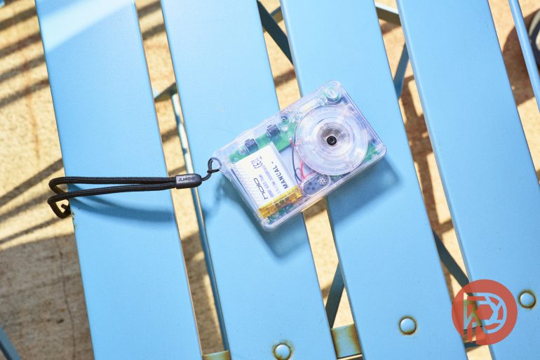 A small, transparent film camera with a wrist strap resting on a bright blue slatted bench.