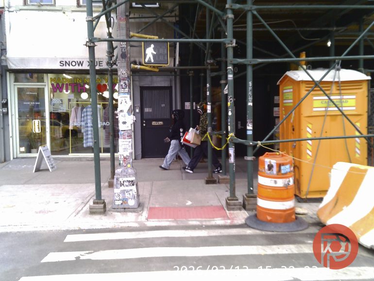 Two people walk under scaffolding past a store and a portable toilet on a city sidewalk.