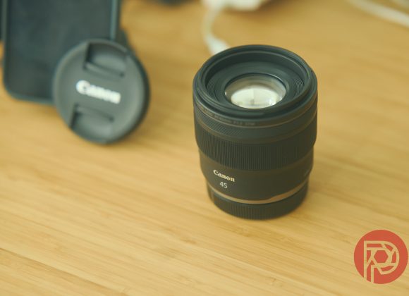 A Canon 45mm camera lens on a wooden surface, with a lens cap and phone in the background.
