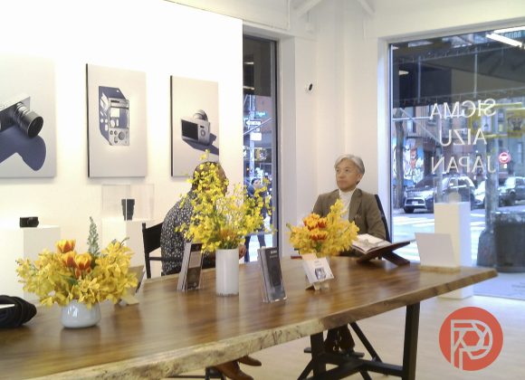 Two people sit at a wooden table with yellow flowers in a modern gallery with photography on the walls.