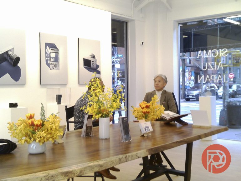 Two people sit at a wooden table with yellow flowers in a modern gallery with photography on the walls.