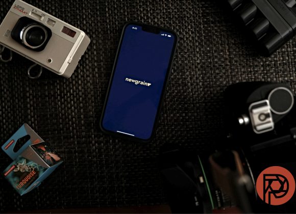 A smartphone with newgrain on screen, surrounded by cameras, film, and camera gear on a dark woven surface.