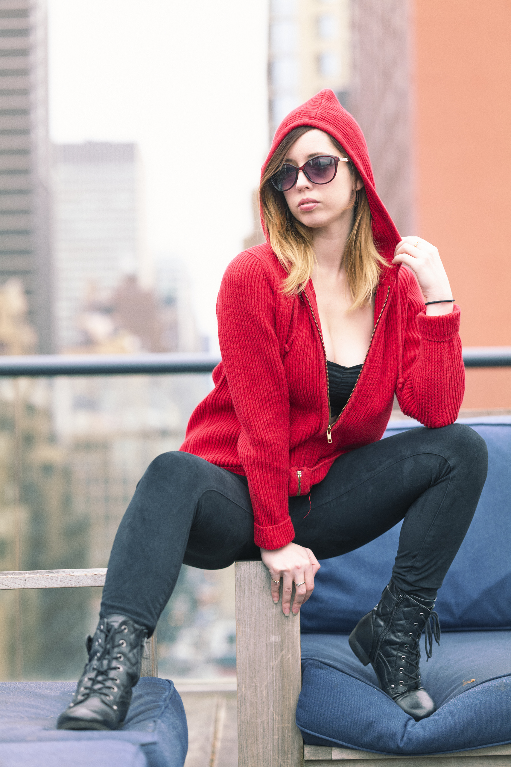 Woman in a red hoodie and sunglasses poses confidently on a rooftop with city buildings in the background.