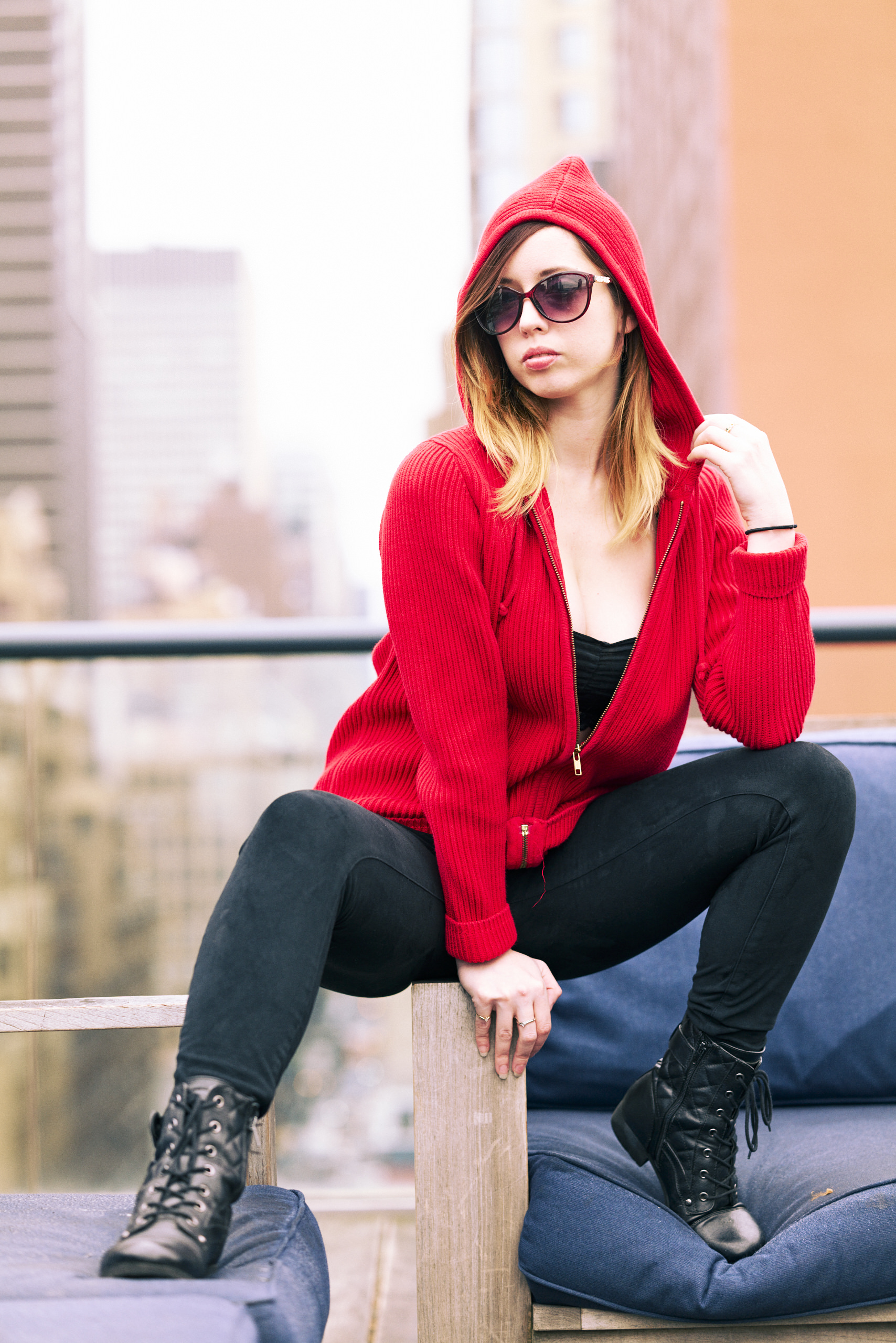 Woman in a red hooded jacket, sunglasses, and black boots poses on a rooftop with city buildings behind her.