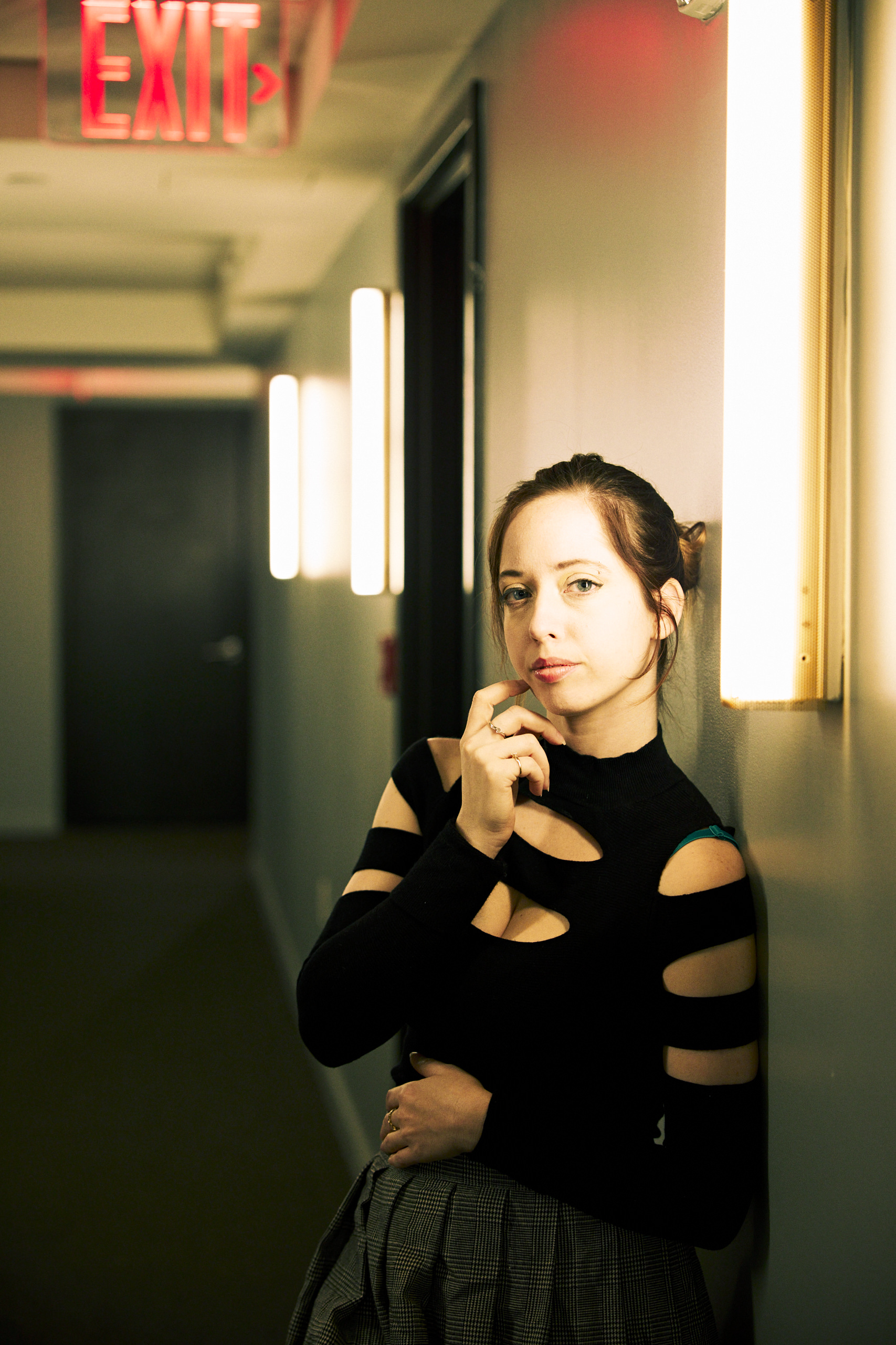 Woman in a cut-out black top leans against a hallway wall under fluorescent lights, with an exit sign above.