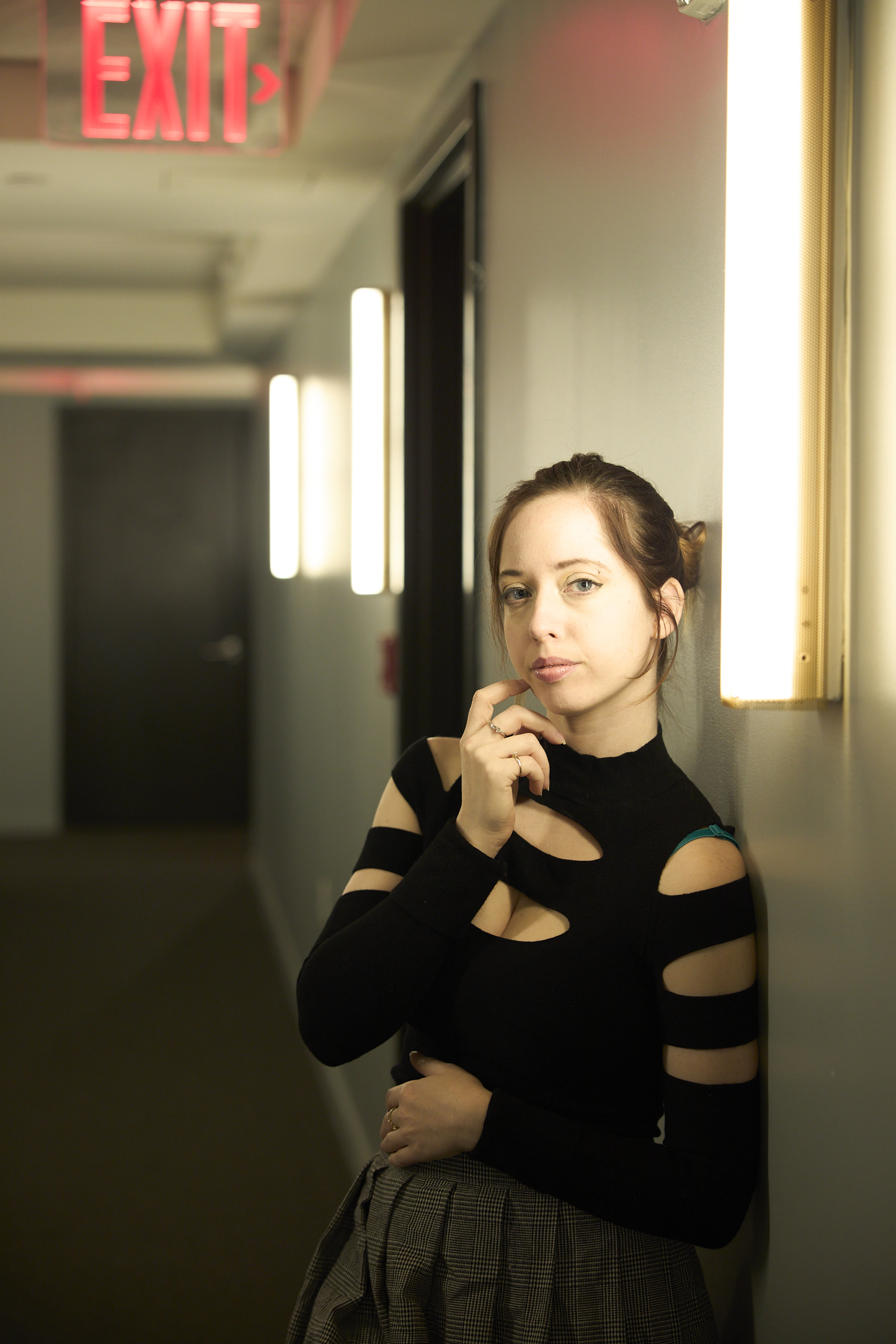 Woman in a black cutout top leans against a hallway wall, illuminated by vertical lights and an exit sign above.