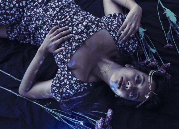 Woman in a floral dress lies on dark fabric with scattered purple flowers, wearing a necklace and headpiece.