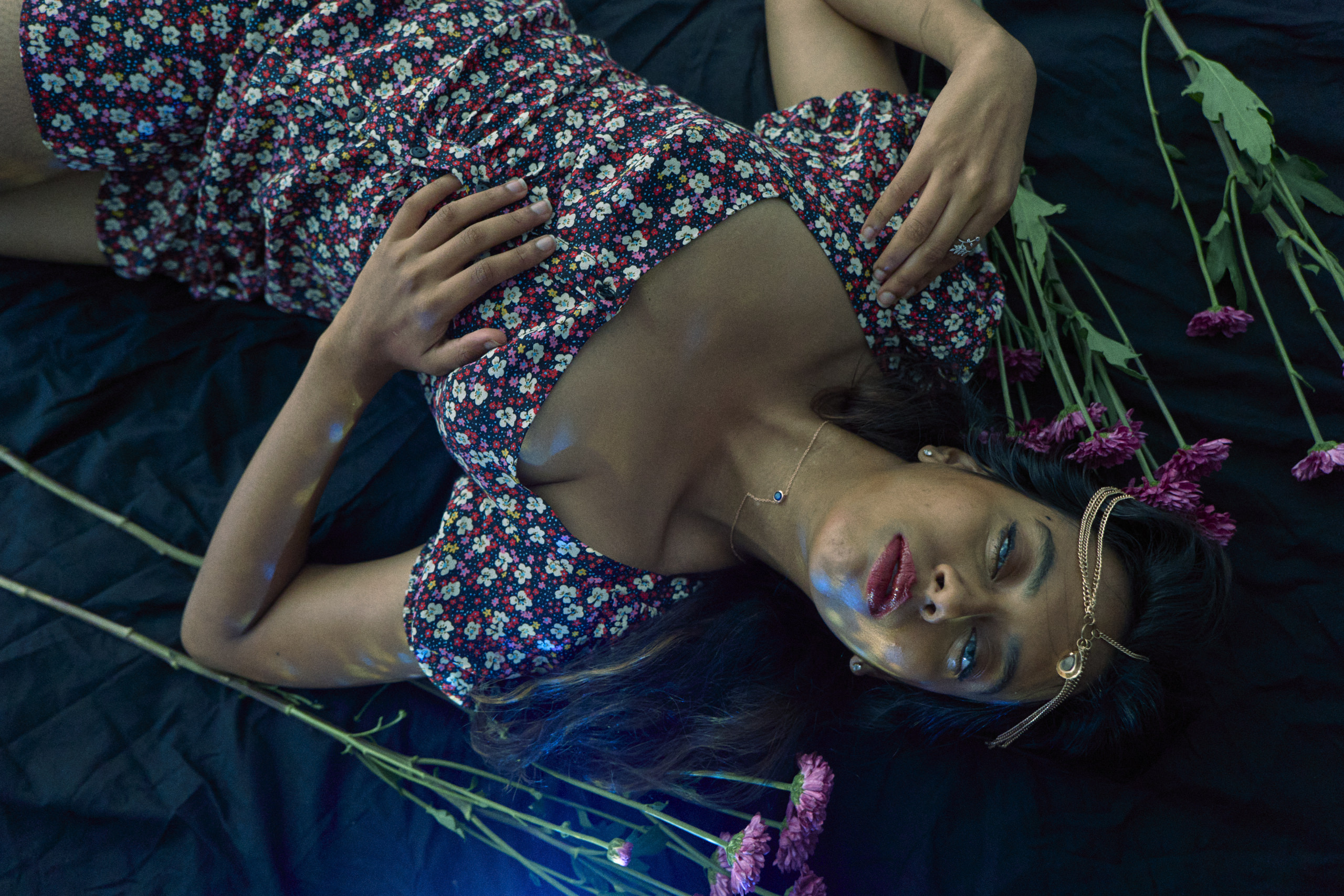 Woman in a floral dress lies on a dark surface, surrounded by pink flowers, gazing upward.