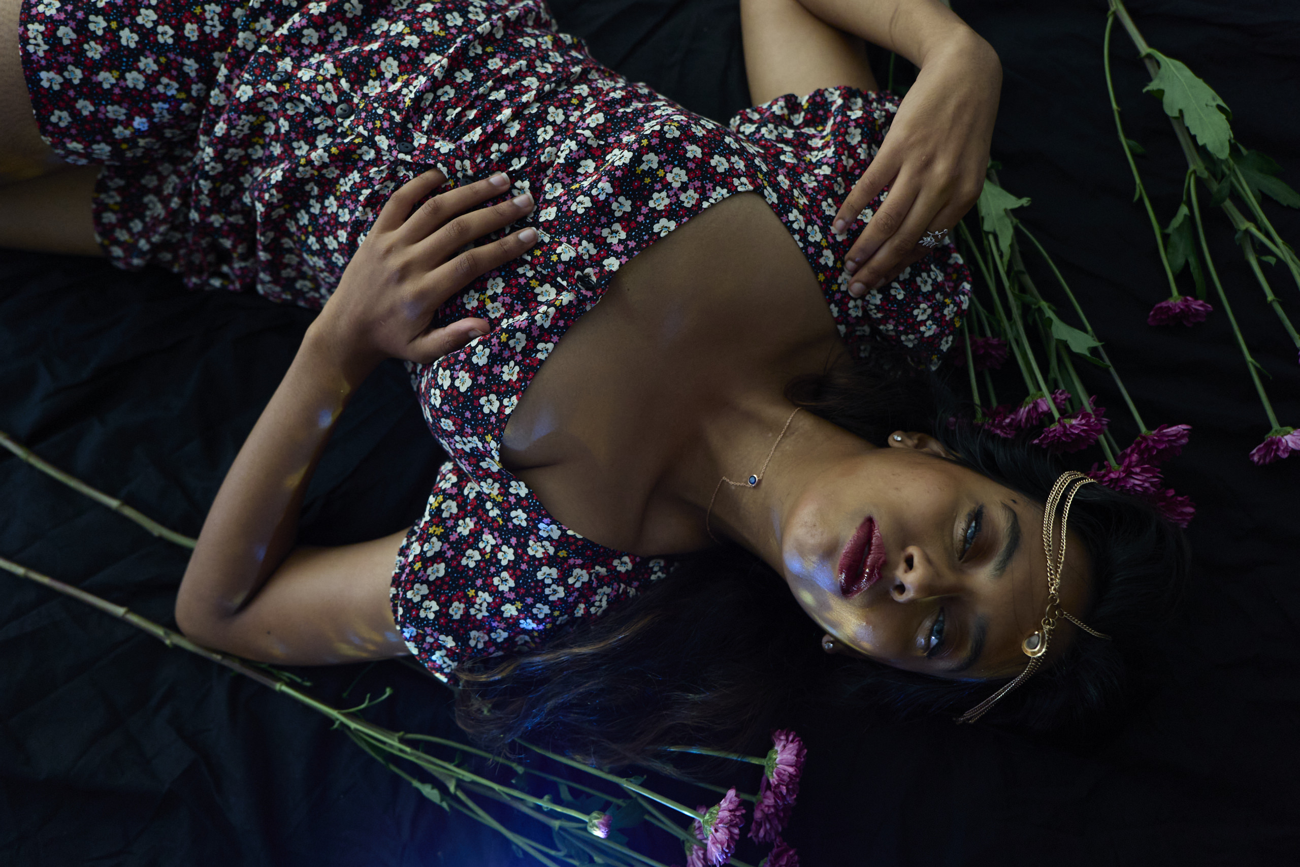 Woman in a floral dress lies on black fabric, surrounded by pink flowers, wearing a gold headpiece and necklace.