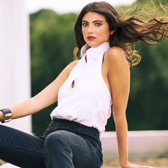 Woman with long brown hair wearing a sleeveless white top and black jeans poses outdoors, hair flowing in the wind.