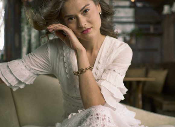 Woman in a white dress sits on a couch indoors, resting her head on her hand and looking at the camera.