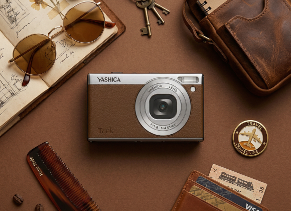 Flat lay of a brown camera, sunglasses, wallet, notebook, and travel-themed items on a brown surface.