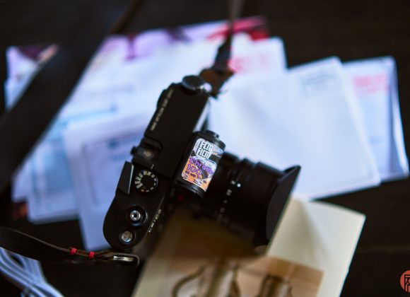 A film camera with a roll of film on top sits on a table with letters and envelopes in the background.