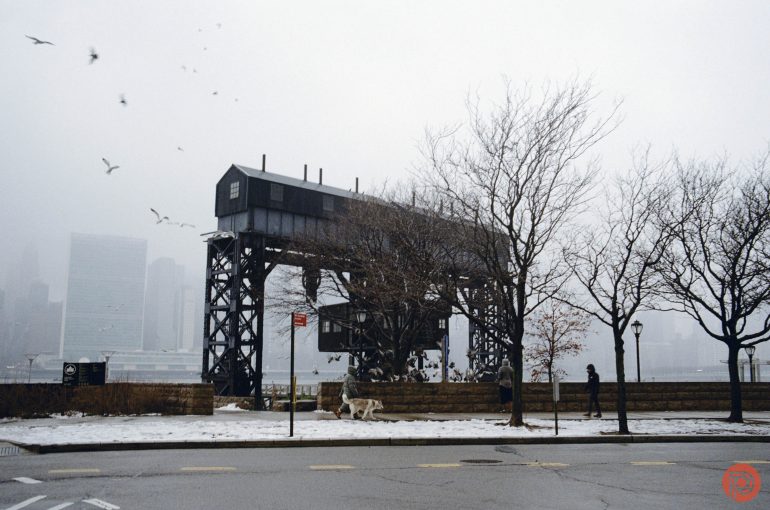 A misty riverside scene with an industrial structure, bare trees, birds flying, and snow on the ground.