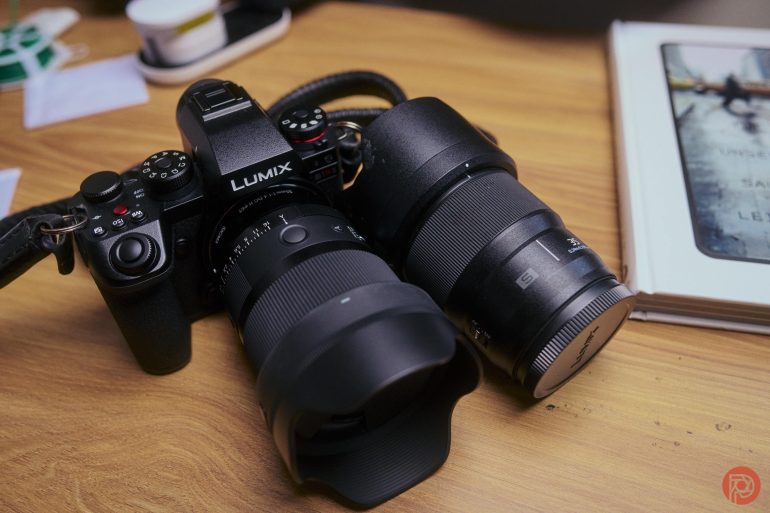 A Lumix camera with two large lenses sits on a wooden desk next to a closed book.