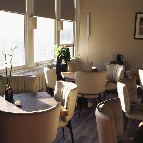 Sunny cafe interior with empty beige chairs, wooden tables, and potted plants by large windows.