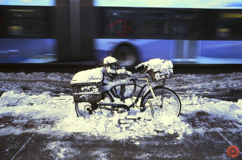 A bicycle covered in snow is parked on a snowy street with a blurred bus passing in the background.