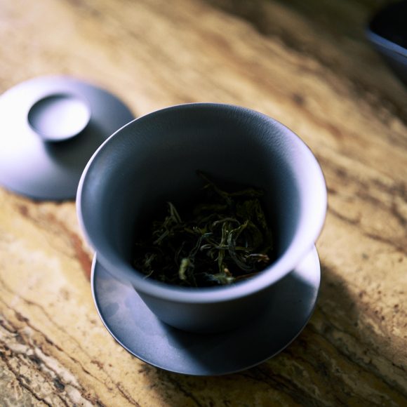 A blue ceramic cup with a lid and saucer holds loose tea leaves on a marbled surface.