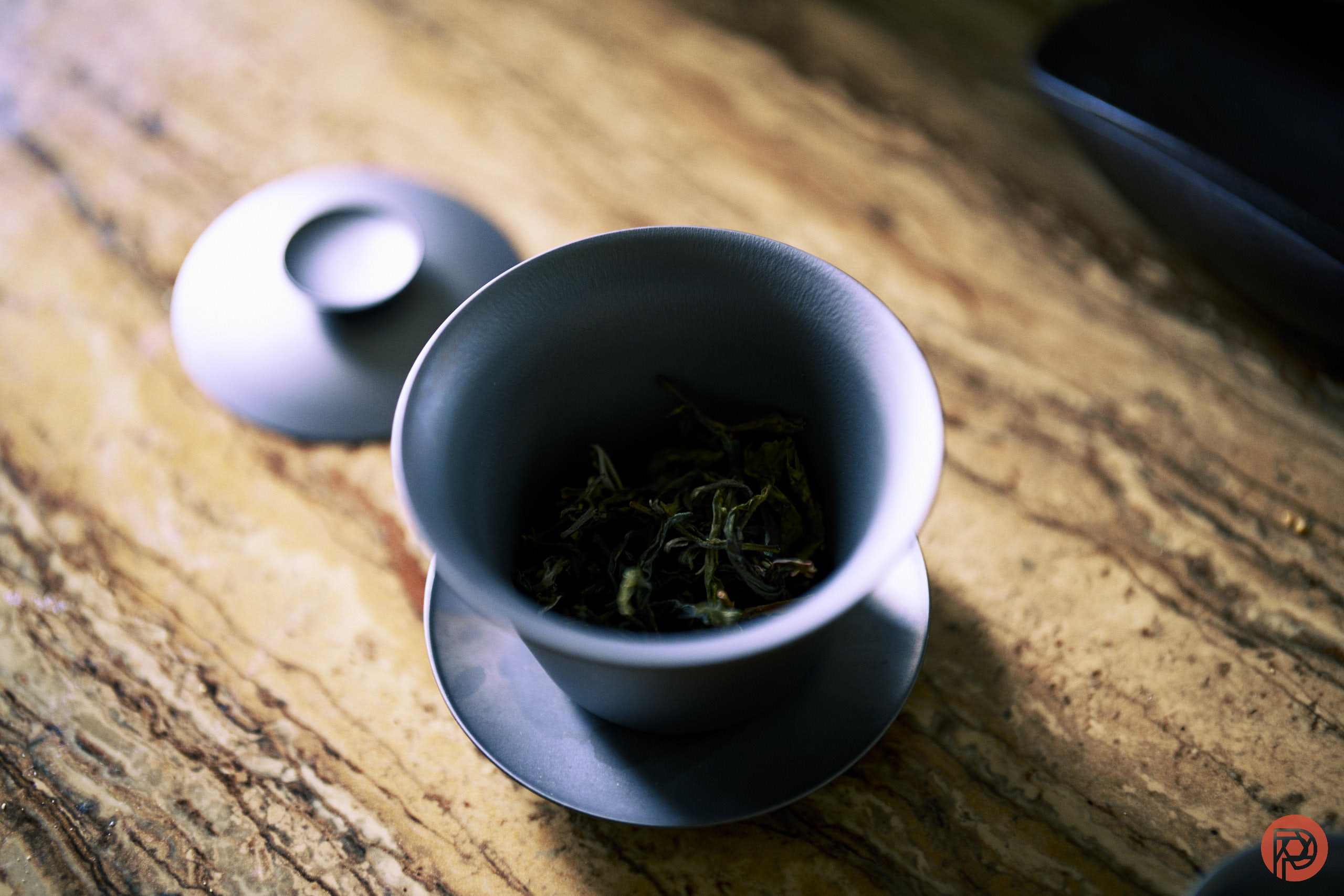Need to Recharge? We Can Help A blue ceramic cup with a lid and saucer holds loose tea leaves on a marbled surface.