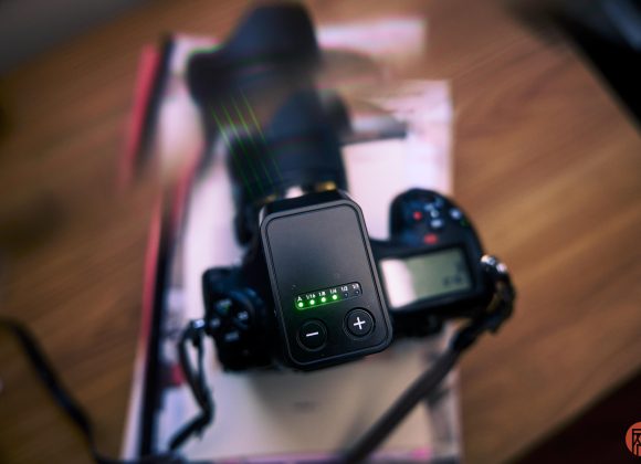 A digital camera with a lens and a control device sits on a wooden table, viewed from above.