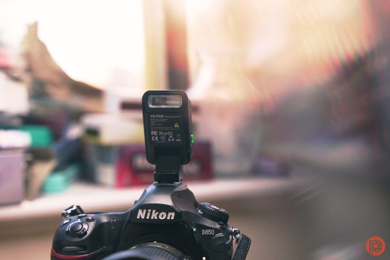 A Nikon D850 camera with an external flash transmitter attached, blurred background.