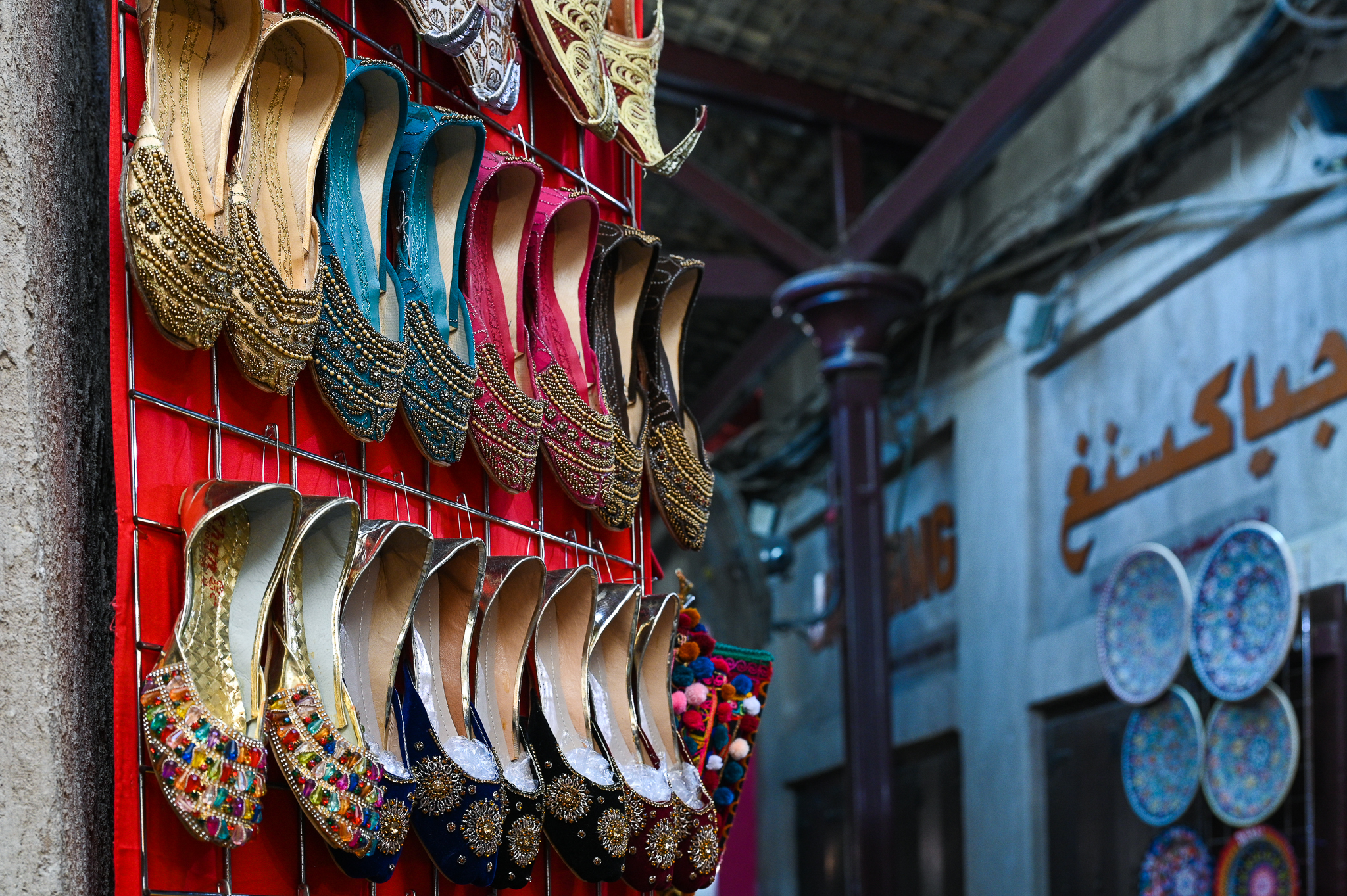 Colorful traditional shoes are displayed on a red rack at an outdoor market with shops in the background.