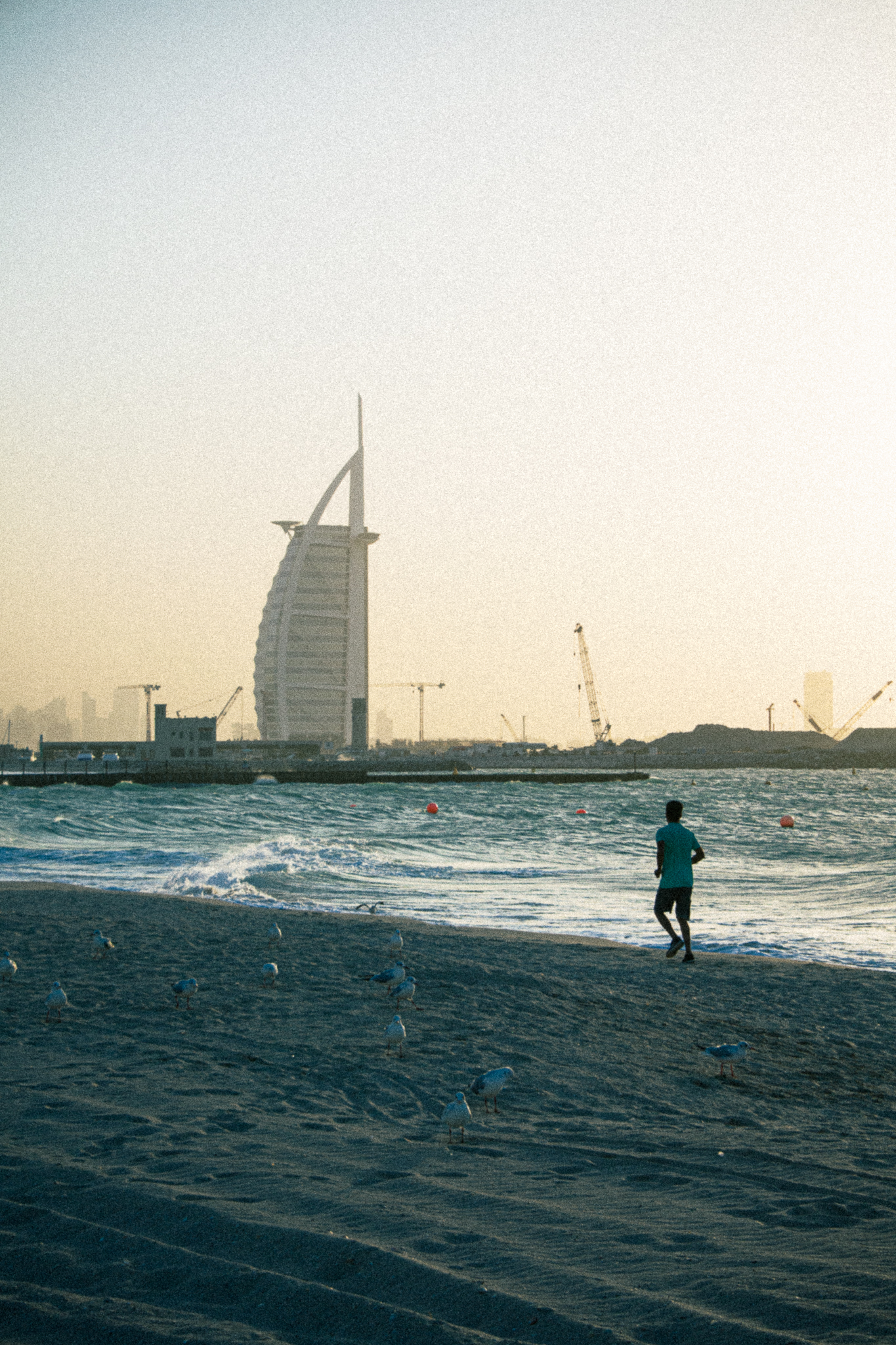 A person walks along a beach with the Burj Al Arab and cranes visible in the background at sunset.