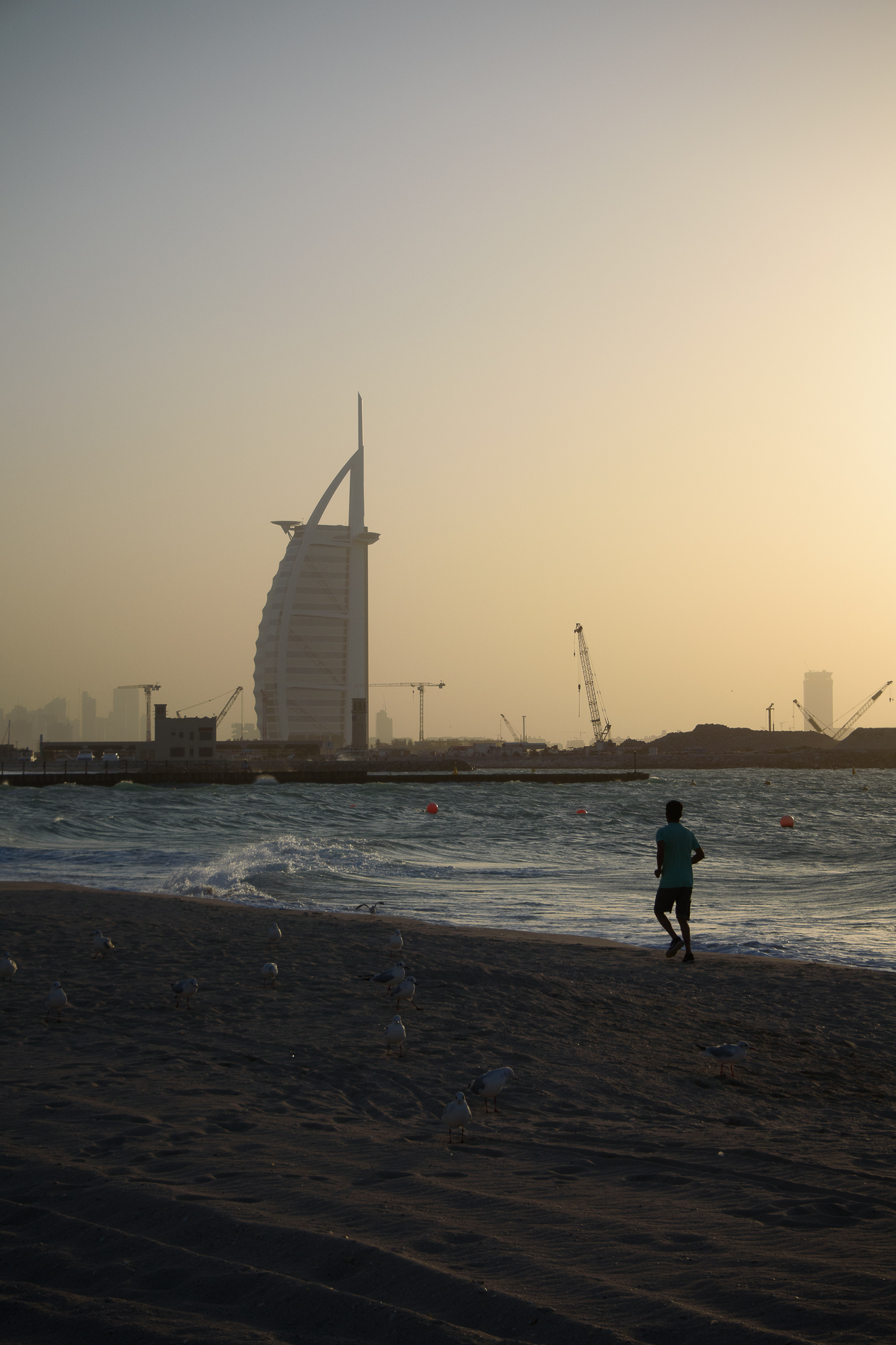 Person walks on a beach at sunset with the Burj Al Arab and cranes visible in the background.