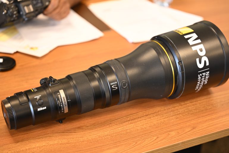 A large Nikon NPS telephoto camera lens lying on a wooden table near some papers.