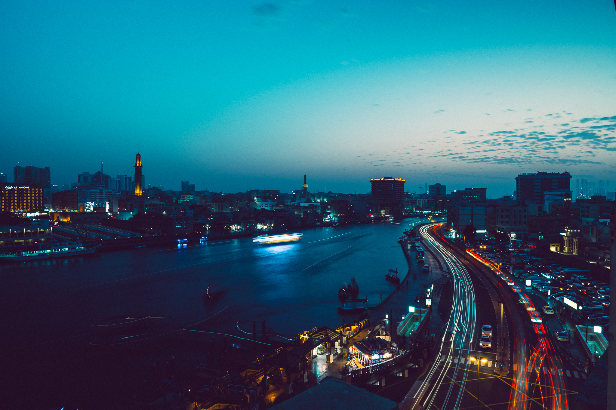 Cityscape at dusk with light trails from cars, illuminated buildings, and a river reflecting the evening sky.