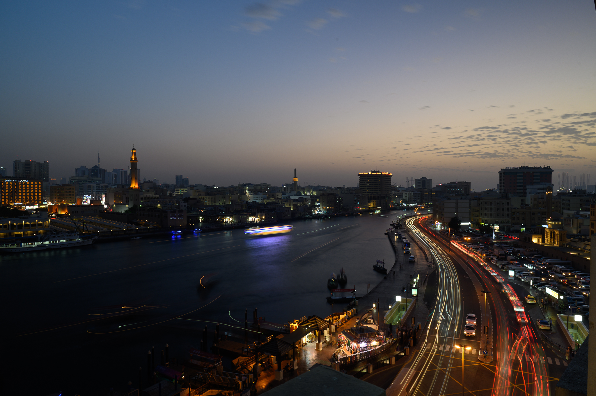 Nighttime cityscape with light trails from cars by a river, buildings illuminated along the waterfront.