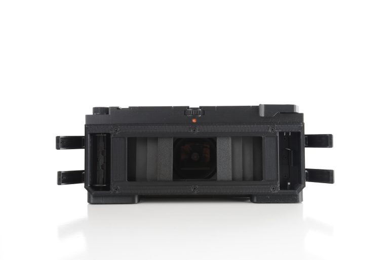 A black film camera with its back open, showing the film compartment and inner mechanisms.