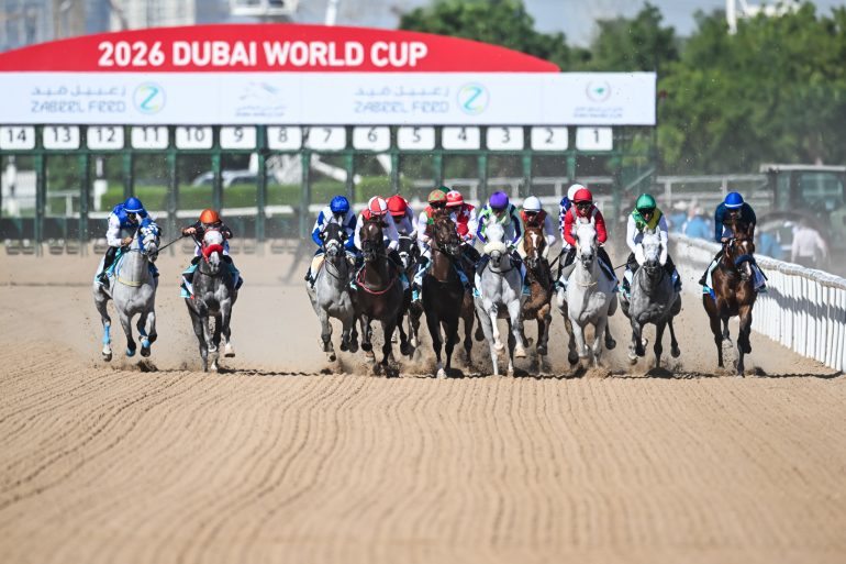 Horses and jockeys racing on a sandy track at the 2026 Dubai World Cup, with the event sign overhead.