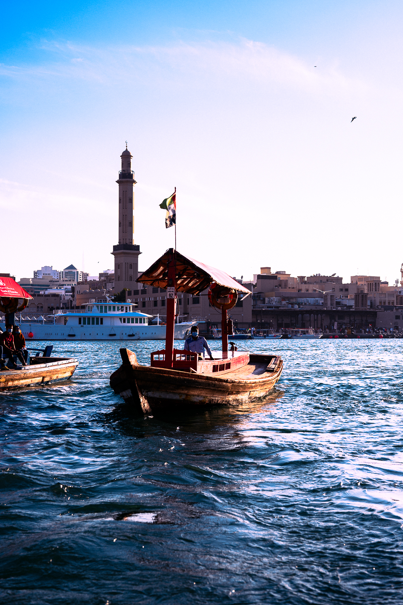 A wooden boat floats on a river with city buildings and a tall minaret in the background under a blue sky.