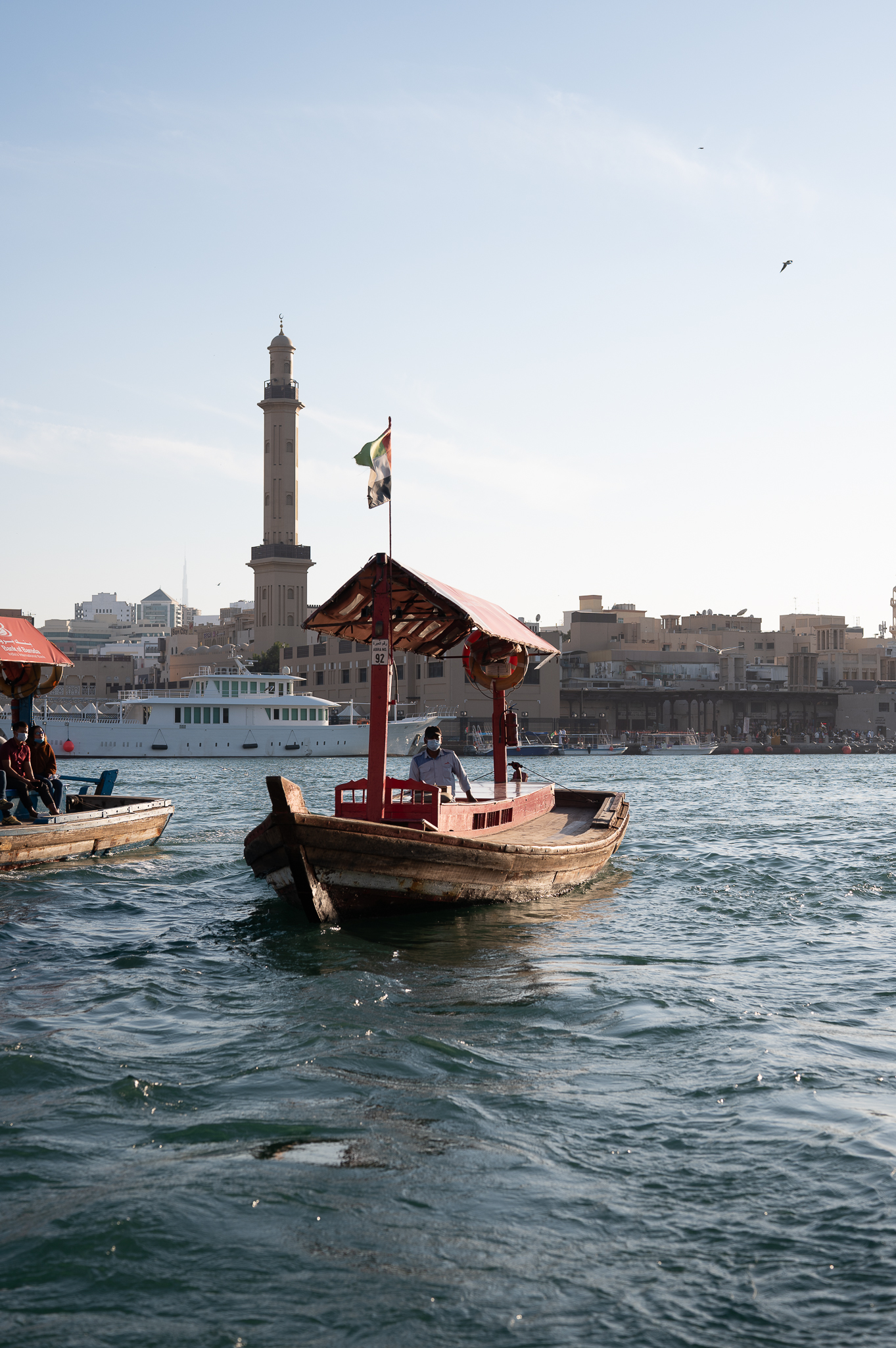 A wooden boat with a canopy floats on water near a city skyline with a tall minaret in the background.