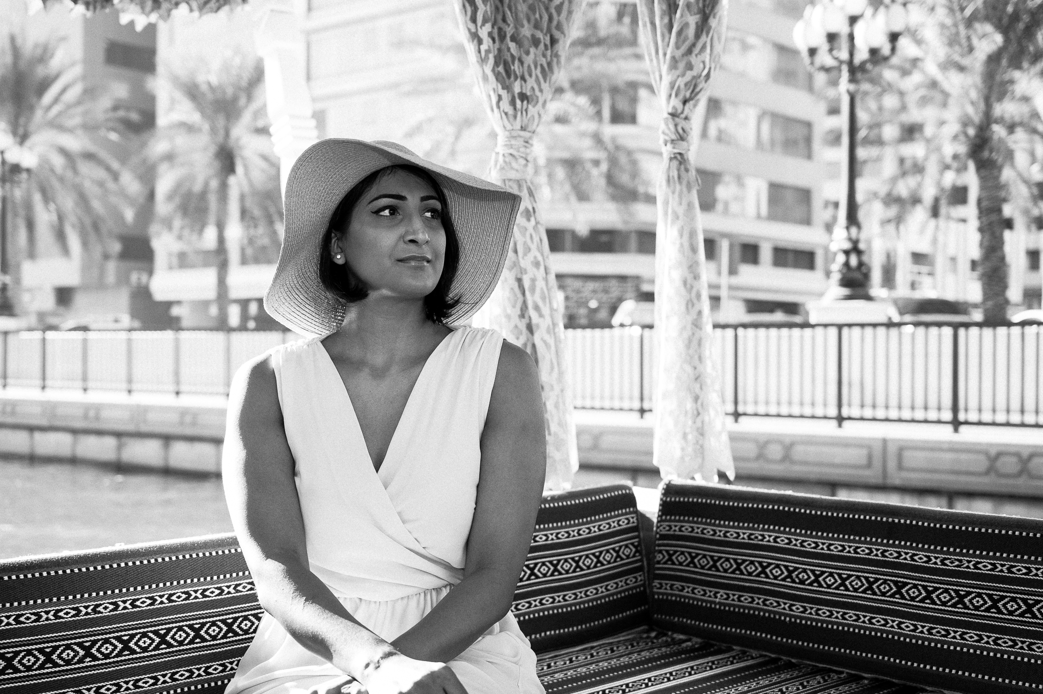 Woman in a sunhat and dress sits on a patterned bench outdoors, city buildings and palm trees in background.