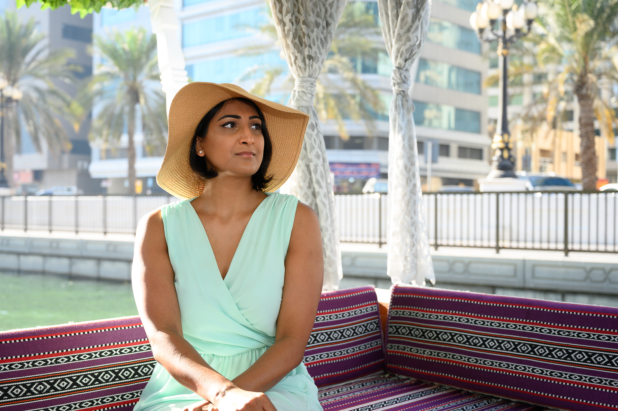 A woman in a wide-brimmed hat sits on a patterned bench outdoors, with palm trees and buildings behind her.