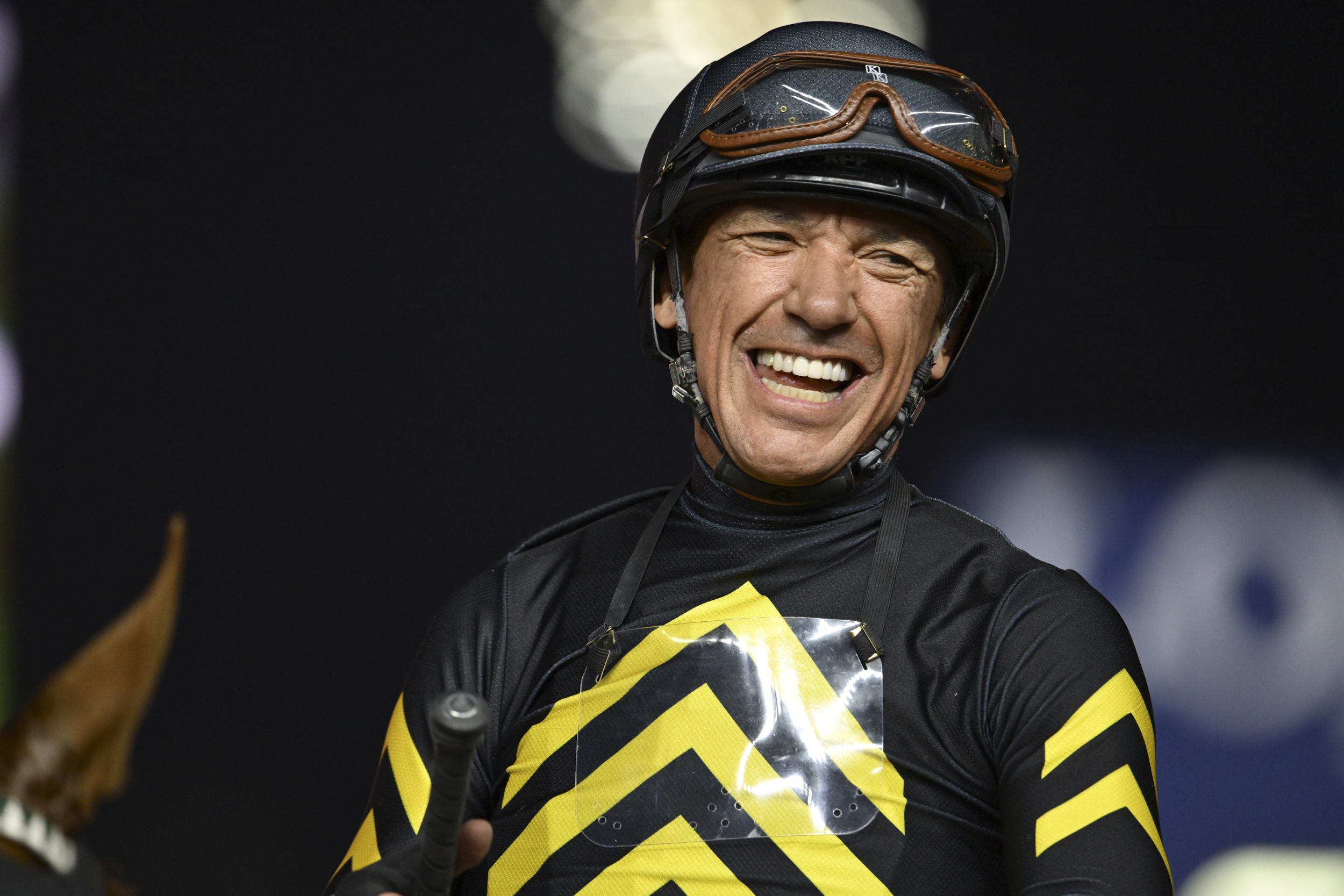 A smiling jockey in black and yellow racing attire and helmet stands holding riding gear.