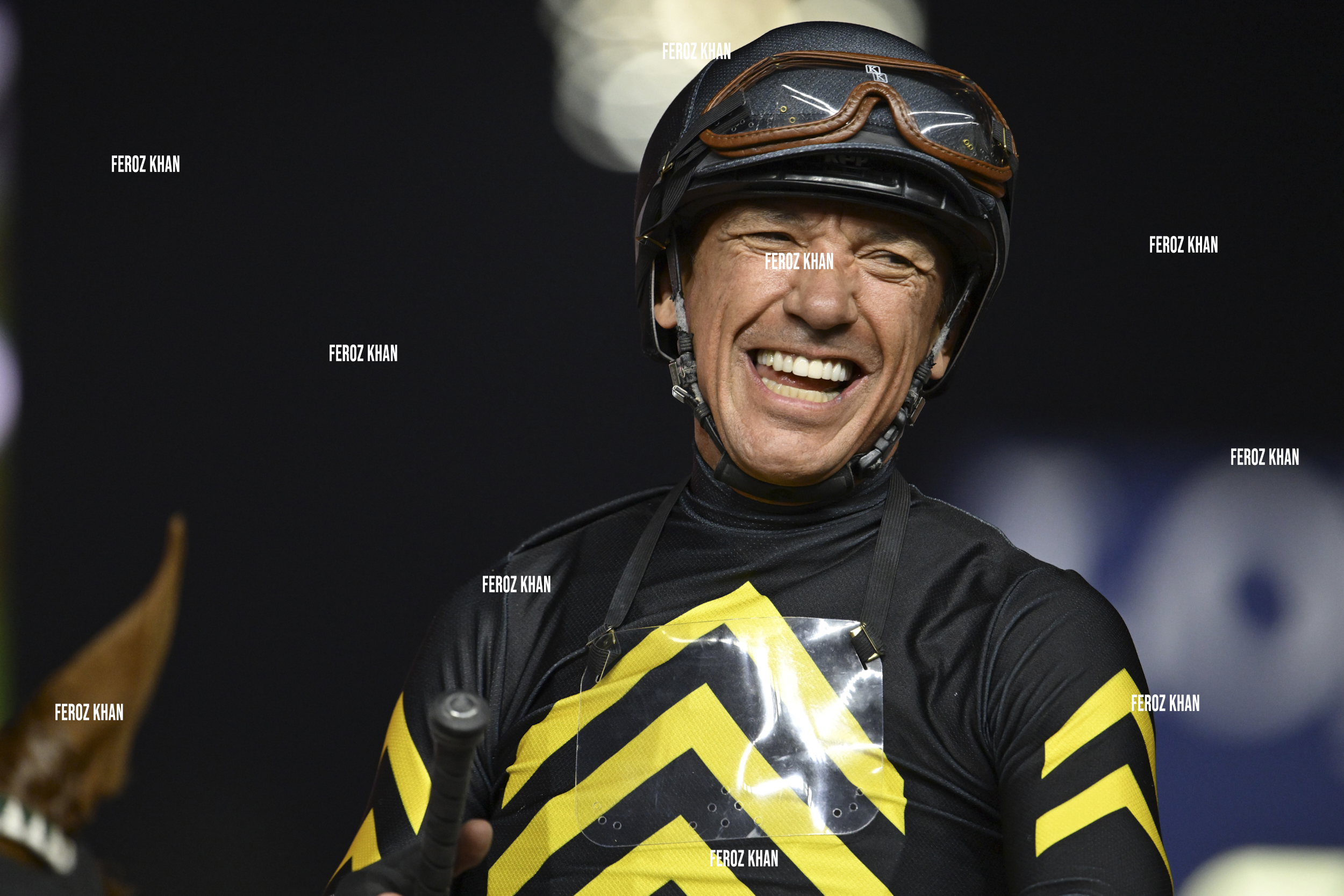 A smiling jockey in black and yellow racing gear and helmet stands holding riding equipment.