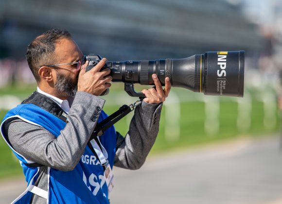A photographer in a blue vest uses a large telephoto camera lens outdoors near a grassy area.
