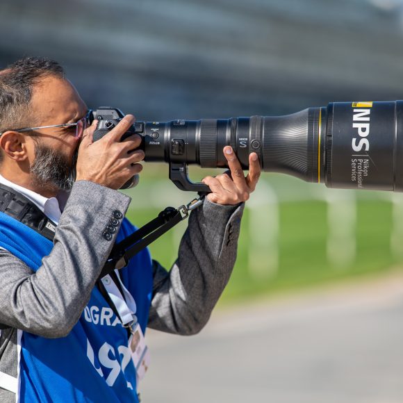 A photographer in a blue vest uses a large telephoto camera lens outdoors near a grassy area.