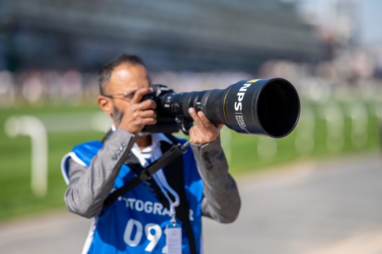 A photographer in a blue vest aims a large camera with a telephoto lens at an outdoor event.