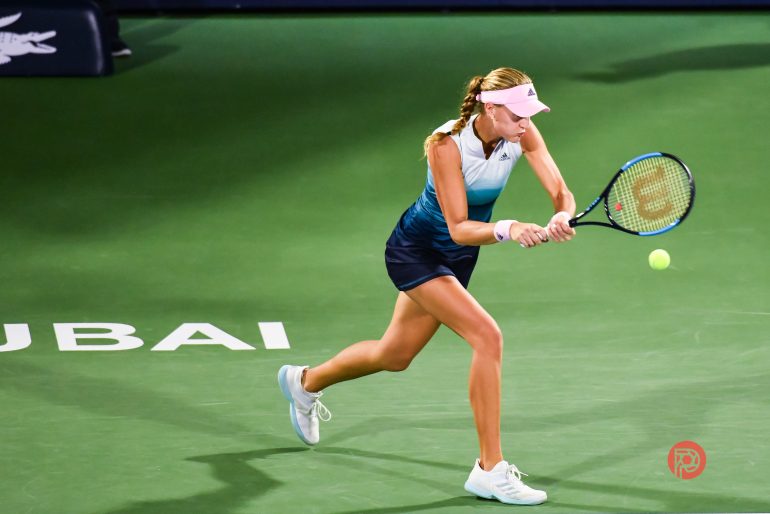 Female tennis player in blue outfit hits a backhand shot on a green court with DUBAI written on it.