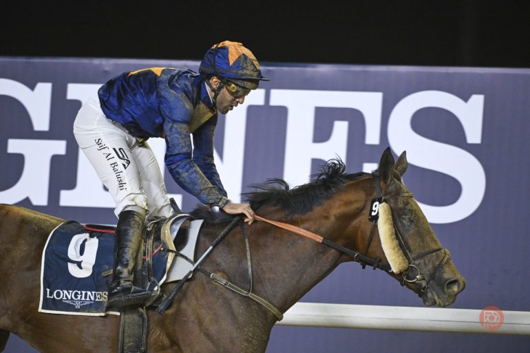 Jockey in blue and orange silks riding horse number 9 during a race, Longines sign in background.