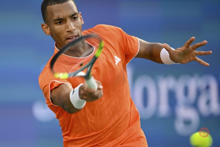 A tennis player in an orange shirt swings a racket to hit a ball during a match.