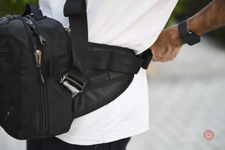 Person wearing a black sling bag and fastening a waist strap outdoors, dressed in a white shirt.